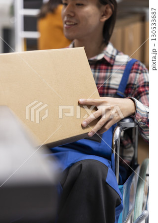 Male worker in wheelchair prepares packages for custom orders and local shipment. Home business logistics center supports chronic impairment with accessible shelving units and packaging station. 130533687