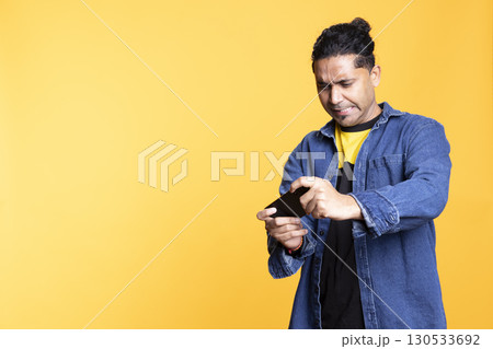 Indian single player having fun with mobile video games against orange background, playing an e sport tournament on smartphone. Young guy with man bun having a modern lifestyle. 130533692