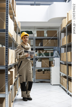 Female worker conducting manual labor surrounded by packages and crates, staff managing shipping, delivery and inventory control tasks. Warehouse interior supply and demand operations. Female worker conducting manual labor surrounded by packages and crates, staff managing shipping, delivery and inventory control tasks. Warehouse interior supply and demand operations. 130534822