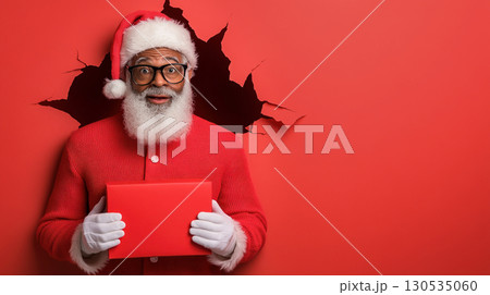 African American Santa, Black Santa Claus with red box his hands looks through torn hole red paper background jumping, points his hand empty place copy space background. 130535060