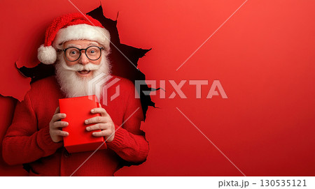 Emotional Santa Claus with red box in his hands looks through torn hole red paper background jumping, points his hand to empty place of copy space background. Xmas copy space 130535121