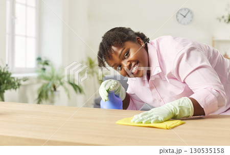 Positive young African American housewife cleaning and wiping desk at home with rag and spray bottle Positive young African American housewife cleaning and wiping desk at home with rag and spray bottle 130535158