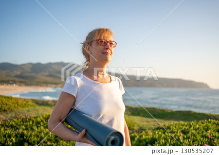 Pensioner woman holding yoga mat standing on ocean Pensioner woman holding yoga mat standing on ocean 130535207