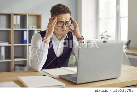 Tired young exhausted overworked business man sitting at the desk on his workplace in office. Tired young exhausted overworked business man sitting at the desk on his workplace in office. 130535793