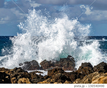 Powerful Ocean Wave Crashing Against Dark Rocks Creating White Foam and Spray Under a Cloudy Blue Sky 130540490