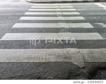 Pedestrian crosswalk painted on asphalt road with visible wear and cracks, located at intersection, with curb and street in background 130540855