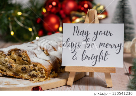 Note with saying MAY YOUR DAYS BE MERRY AND BRIGHT congratulations. Traditional German Christmas stollen cake in background of handmade festive decor. Winter holiday treats. Vegan baking recipe 130541230