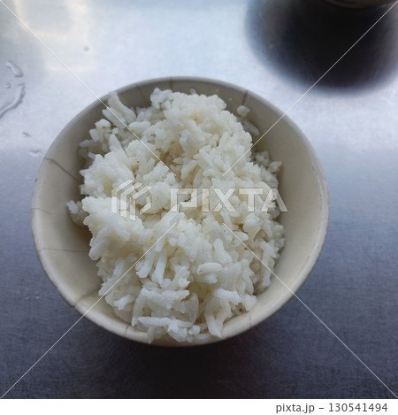 Bowl of cooked white rice on metallic surface with dark container in background, showing simple and clean presentation, evoking sense of freshness and readiness to eat Bowl of cooked white rice on metallic surface with dark container in background, showing simple and clean presentation, evoking sense of freshness and readiness to eat 130541494