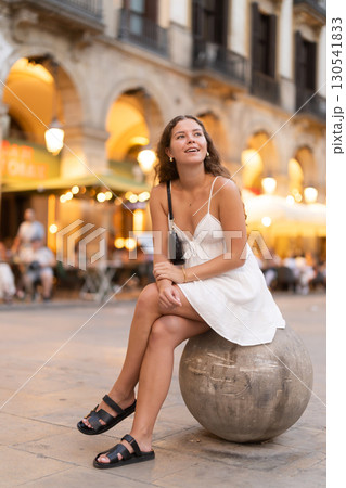 Evening Royal square in Barcelona, girl in sundress is sitting on stone ball Evening Royal square in Barcelona, girl in sundress is sitting on stone ball 130541833