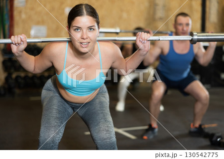Energetic young female athlete practicing sit-ups with rod near other people gym 130542775