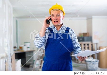 Worker in a protective helmet and blue overall discussing a repair plan on mobile phone Worker in a protective helmet and blue overall discussing a repair plan on mobile phone 130543147