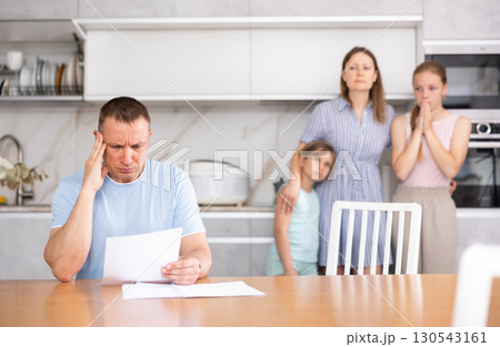 Disgruntled father carefully reads documents. The whole family stands in fear in background Disgruntled father carefully reads documents. The whole family stands in fear in background 130543161