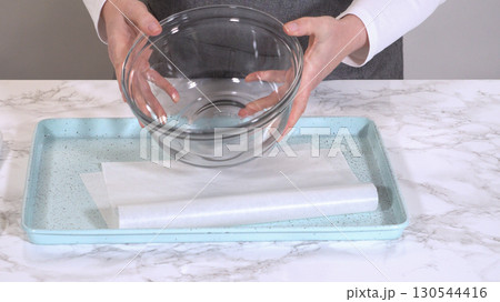 A person in a gray apron holds a clear glass mixing bowl above a blue speckled kitchen tray lined with parchment paper on a marble counter. The scene is ready for food preparation. A person in a gray apron holds a clear glass mixing bowl above a blue speckled kitchen tray lined with parchment paper on a marble counter. The scene is ready for food preparation. 130544416