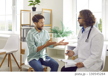 Young man patient talking to a male doctor in his office in clinic during medical examination. Young man patient talking to a male doctor in his office in clinic during medical examination. 130545264