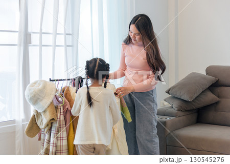 On a bright winter day a woman helps a girl pick out warm clothing from a rack, preparing for the cold weather, the outfits suggesting a season with winter hats and coats. 130545276