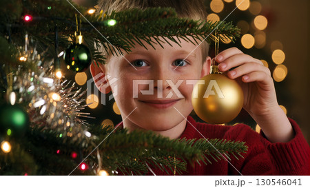 Christmas boy holding golden bauble near decorated tree with lights and ornaments, joyful holiday preparation moment 130546041