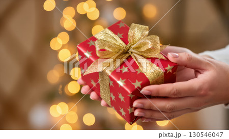 Christmas gift in hands with red wrapping paper, gold stars and glitter ribbon, festive bokeh lights in background 130546047