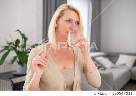 Middle aged woman holding pill between fingers and glass of water, focused expression, sitting in cozy living room  130547031