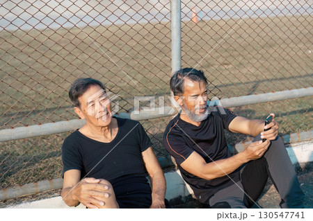 Content elderly men relaxing together and using a smartphone after a workout on the track. Emphasizing friendship, wellness, and active aging. 130547741
