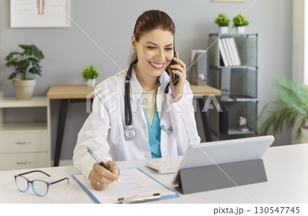 Doctor sitting at the desk in office talking by mobile phone and making notes consulting patients Doctor sitting at the desk in office talking by mobile phone and making notes consulting patients 130547745