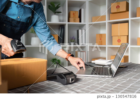 E-commerce entrepreneur using a barcode scanner and laptop to manage inventory and online sales in a modern workspace. 130547816