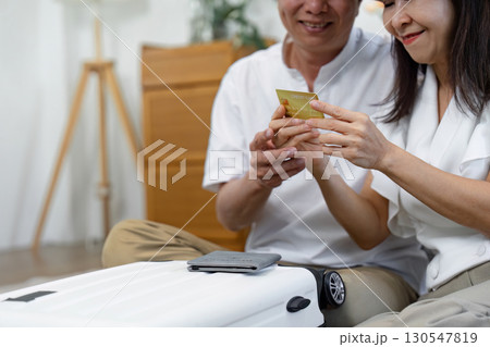 Senior couple planning their travel budget together while holding a credit card and sitting beside a suitcase at home. 130547819