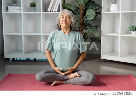Senior woman meditating on a yoga mat at home, promoting wellness and mindfulness. 130547820
