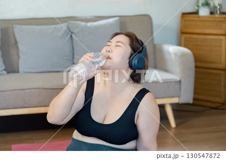 Hydrated woman enjoying water after workout while wearing headphones at home. 130547872