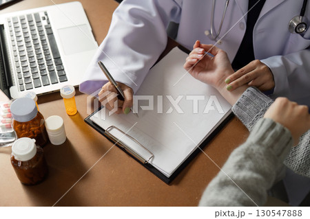 Detail-oriented Muslim female doctor documenting patient information while checking pulse in a modern healthcare office, emphasizing diversity. 130547888