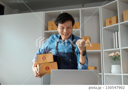 Cheerful Asian male entrepreneur celebrating online sales success while holding packages in a modern workspace. Cheerful Asian male entrepreneur celebrating online sales success while holding packages in a modern workspace. 130547902