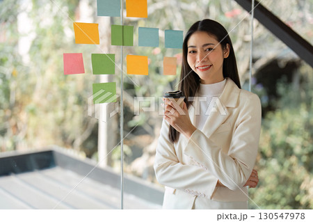 Confident Asian businesswoman holding coffee cup while standing near colorful sticky notes in a modern office. Confident Asian businesswoman holding coffee cup while standing near colorful sticky notes in a modern office. 130547978