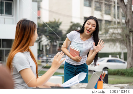 Diverse female student greeting friend while holding notebooks outdoors. 130548348