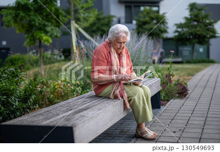 Elderly woman reading a book outdoors in urban garden. Elderly woman reading a book outdoors in urban garden. 130549693