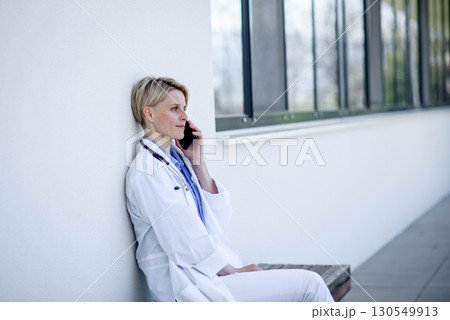 Young female doctor taking a break, sitting on bench in front of hospital and making call. Young female doctor taking a break, sitting on bench in front of hospital and making call. 130549913