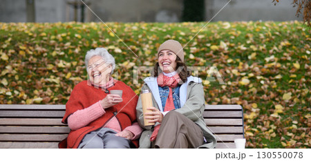 Portrait of a granddaughter and grandmother sitting on bench in autumn park, drinking coffee and laughing together. 130550078