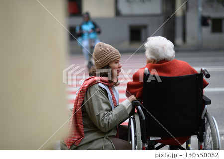 Granddaughter on an autumn walk in the city with her grandmother, pushing her in wheelchair. Rear view. Granddaughter on an autumn walk in the city with her grandmother, pushing her in wheelchair. Rear view. 130550082