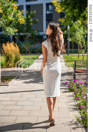 Young woman wearing a white midi dress walking outdoors in Old Money style. Concept of timeless fashion, graceful femininity and understated elegance in modern lifestyle. Young woman wearing a white midi dress walking outdoors in Old Money style. Concept of timeless fashion, graceful femininity and understated elegance in modern lifestyle. 130550508