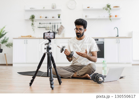 Adult man, sitting on floor in home kitchen, records blog video on fitness topics. Explains exercise methods, holding dumbbells to illustrate. 130550617