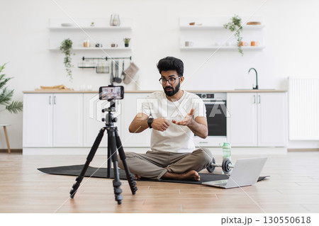 Caucasian adult man in casual outfit recording sports-related vlog discussing smart fitness devices. Room setting is a modern kitchen with exercise equipment visible. 130550618