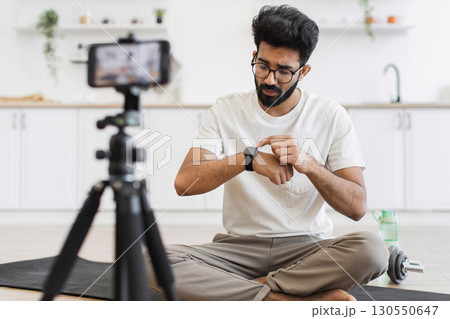 Young adult male recording video blog about smartwatch capabilities for fitness and lifestyle. Scene depicts indoor environment, tripod-mounted smartphone camera, casual setup. Young adult male recording video blog about smartwatch capabilities for fitness and lifestyle. Scene depicts indoor environment, tripod-mounted smartphone camera, casual setup. 130550647