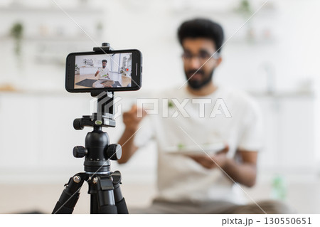 Young adult man sits in kitchen recording video about healthy eating habits. Scene shows him eating and using smartphone camera for vlog, promoting balanced diet and modern lifestyle. 130550651