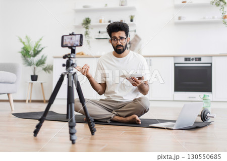 Young adult man sitting on mat in kitchen recording video about healthy eating lifestyle showing food plate to camera. 130550685