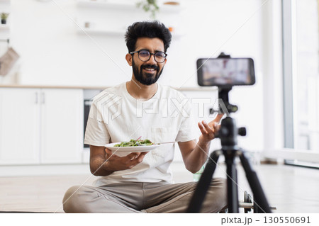 Young adult male with beard records video blog in home kitchen holding salad bowl, discussing healthy eating habits and sharing tips. Scene shows content creation, cooking, modern lifestyle concepts. Young adult male with beard records video blog in home kitchen holding salad bowl, discussing healthy eating habits and sharing tips. Scene shows content creation, cooking, modern lifestyle concepts. 130550691
