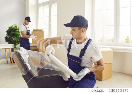 Young male mover packing furniture with plastic wrap in the living room. Young male mover packing furniture with plastic wrap in the living room. 130553173