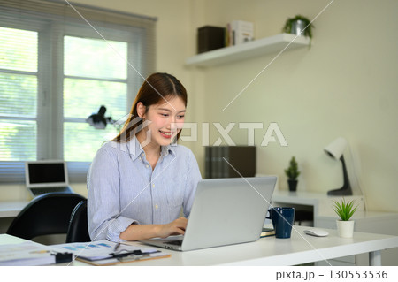 Young woman smiling while working on laptop at home office desk with coffee cup 130553536