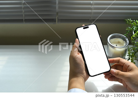 Hand holding smartphone with blank white screen, minimal desk setup with candle and plant. Hand holding smartphone with blank white screen, minimal desk setup with candle and plant. 130553544
