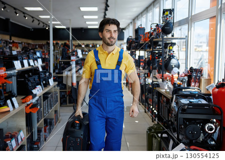 A man dressed in blue overalls is carrying a vacuum cleaner inside a store A man dressed in blue overalls is carrying a vacuum cleaner inside a store 130554125