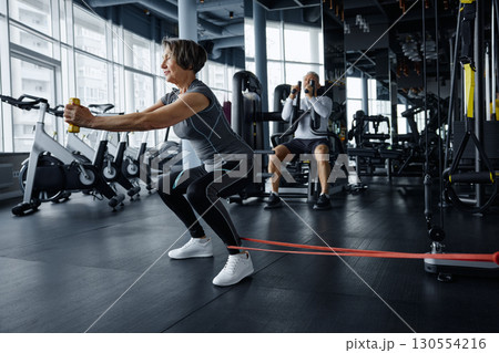 Mature woman doing squats with a rubber band with exercising man on the background Mature woman doing squats with a rubber band with exercising man on the background 130554216