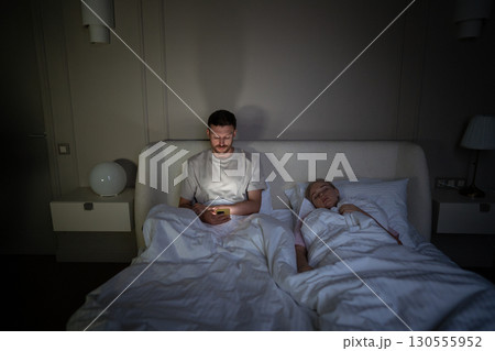 Stressed man scrolls smartphone at night, disturbing sleeping wife, struggling with insomnia 130555952