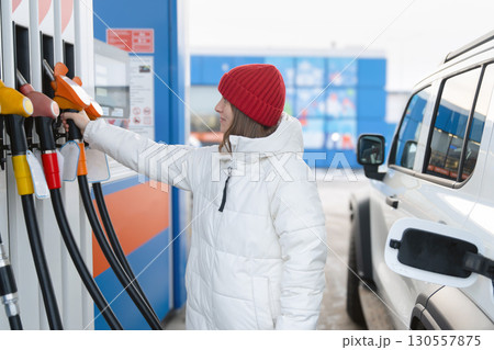 A woman in a white jacket takes off a fuel dispenser gun at a gas station. She was going to fill up her car with gasoline. Gasoline and diesel prices at gas stations. A woman in a white jacket takes off a fuel dispenser gun at a gas station. She was going to fill up her car with gasoline. Gasoline and diesel prices at gas stations. 130557875